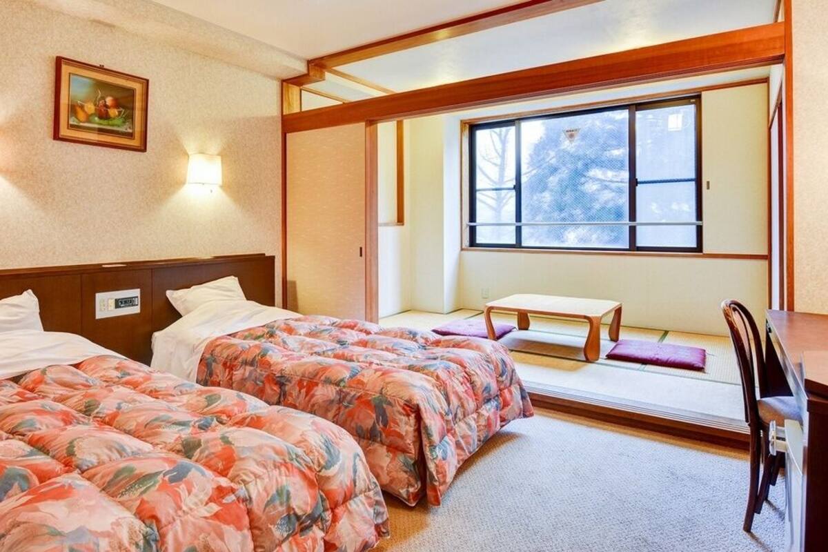 Ein Hotelzimmer mit zwei Betten, einem kleinen Tisch, einem Stuhl und einem Fenster mit Blick auf Bäume.