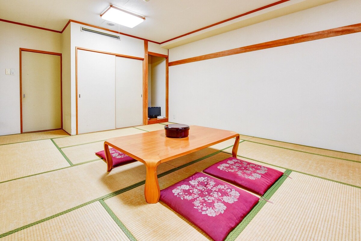 Ein Raum mit Tatami-Matten, einem Holztisch, Kissen, einer Schiebetür und einer Deckenleuchte.