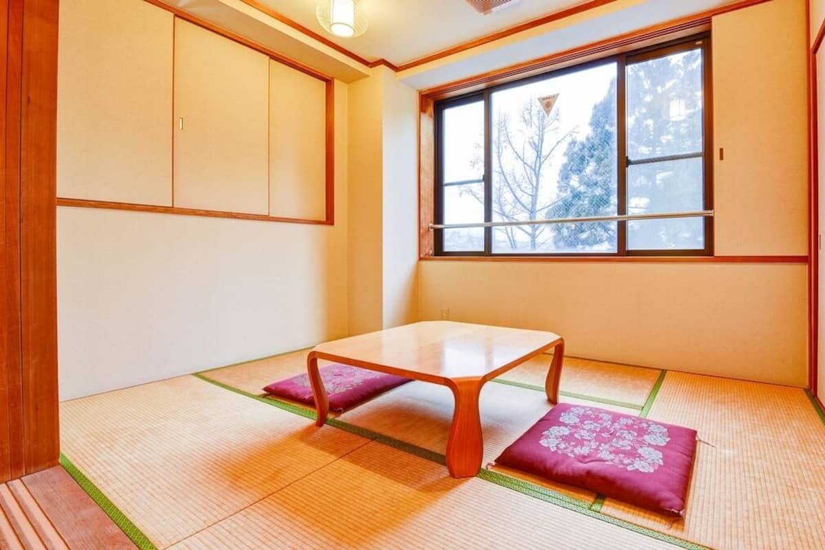 Ein Raum mit Tatami-Matten, einem niedrigen Tisch, Kissen, einem Fenster mit Blick auf Bäume und Schiebetüren.