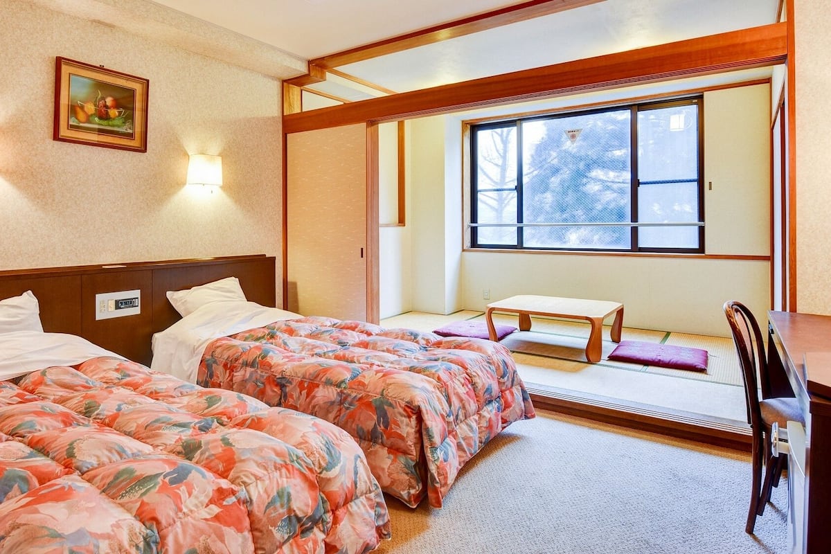 Ein Hotelzimmer mit zwei Betten, einem kleinen Tisch, einem Stuhl und Blick ins Freie durch ein großes Fenster.