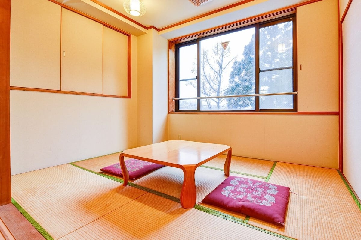 Ein traditioneller japanischer Raum mit Tatami-Boden, einem niedrigen Holztisch und einem Fenster mit Blick auf Bäume.