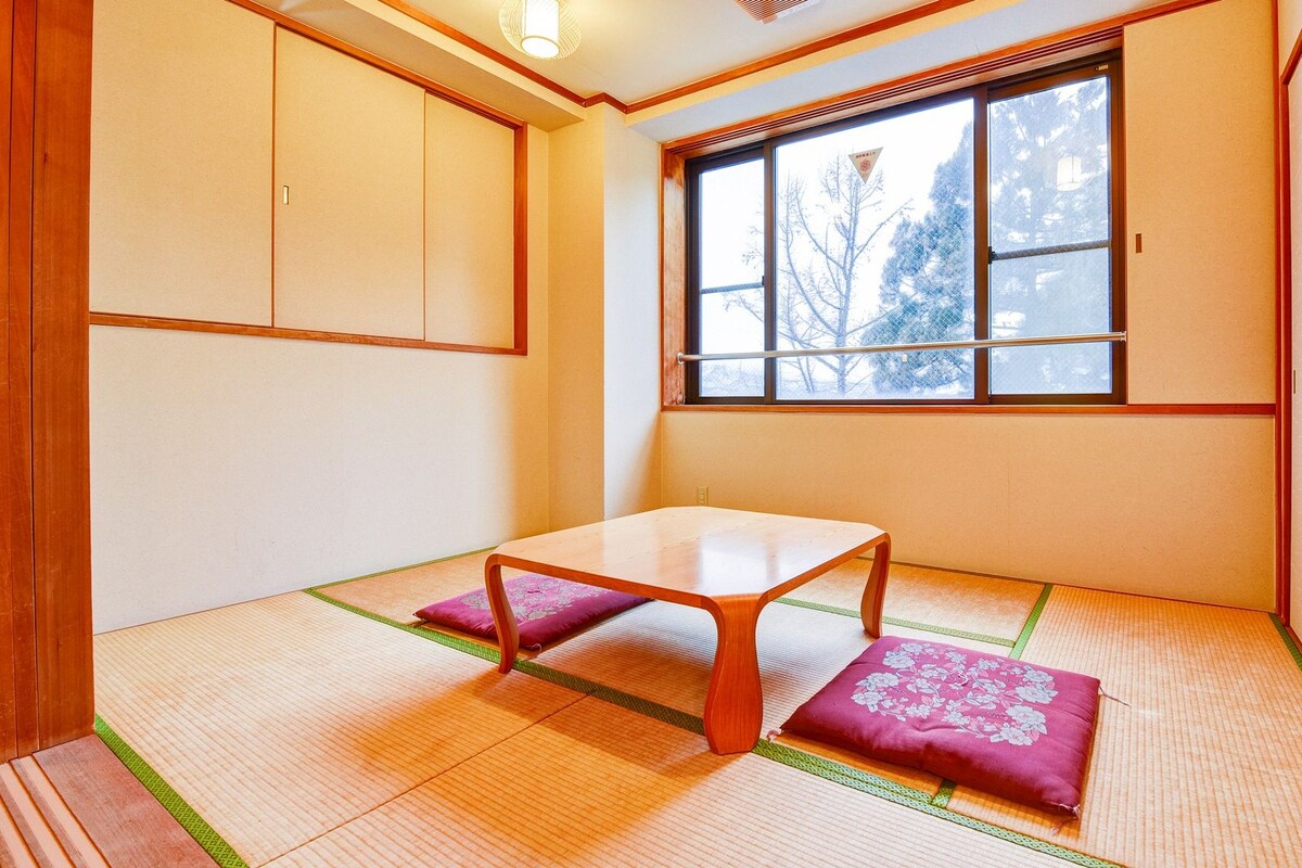 Ein traditioneller japanischer Raum mit Tatami-Boden, einem niedrigen Holztisch und einem Fenster mit Blick auf Bäume.