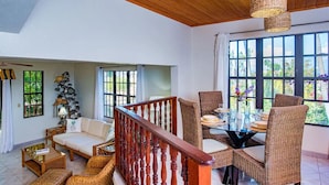 Een ruime woonkamer met een houten plafond, een eettafel met rieten stoelen, en een trap met houten leuning.