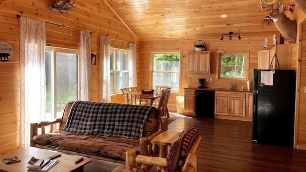 A cabin interior with a sofa, dining table, chairs, a kitchen area, and a refrigerator. - Camp Adirondack at Big Moose Lake (Eagle Bay)
