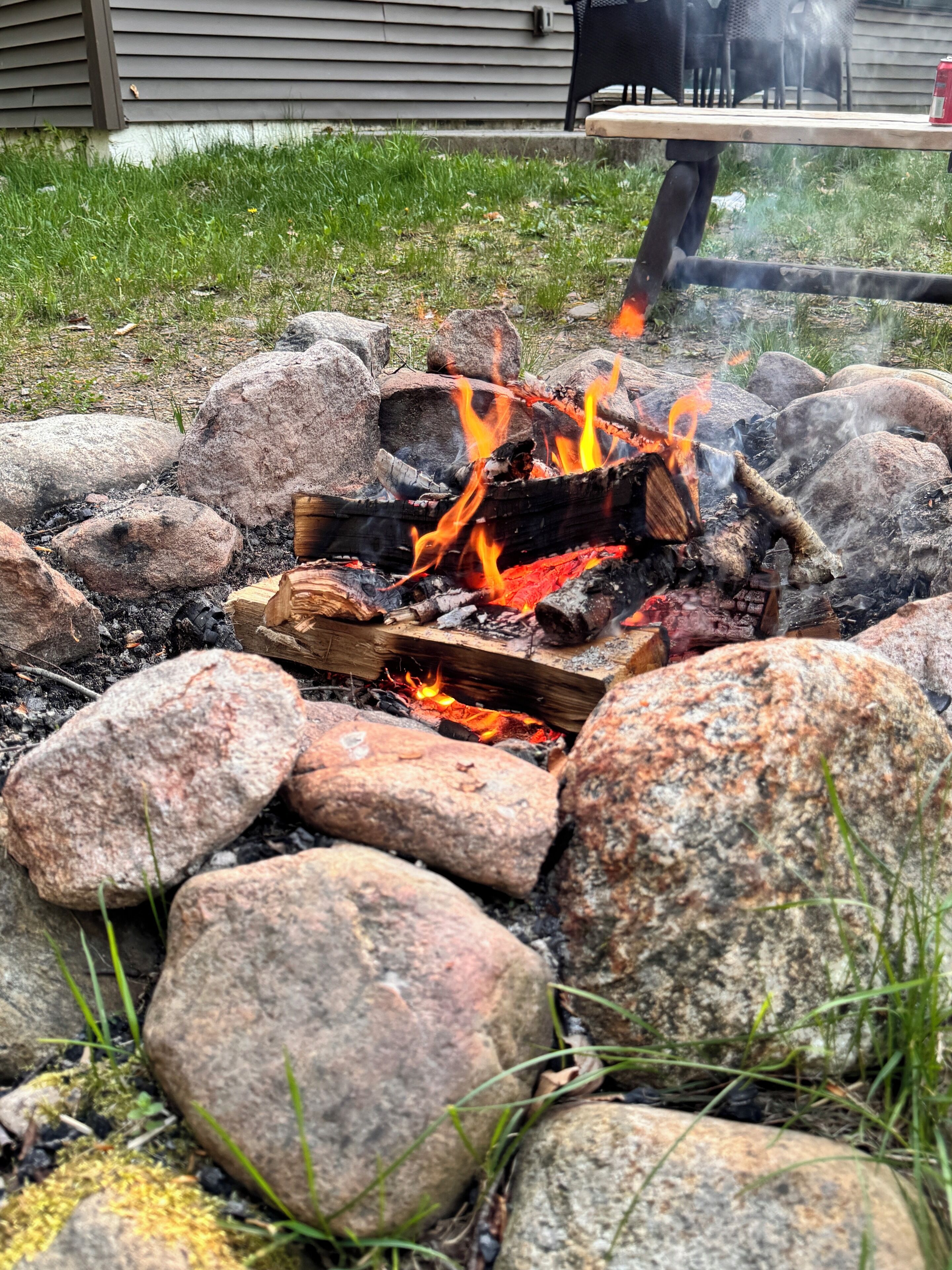 Ein Lagerfeuer mit Holzscheiten und Steinen, umgeben von Gras, mit einem Haus im Hintergrund.
