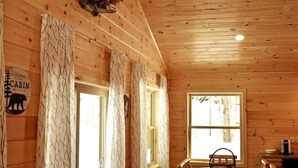 A cabin interior with wooden walls, a dining area, and a window with a view of trees. - Camp Adirondack at Big Moose Lake (Eagle Bay)
