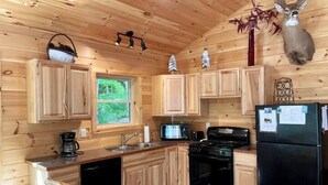 A wooden cabin kitchen with wooden cabinets, a stove, a refrigerator, and a window overlooking greenery. - Camp Adirondack at Big Moose Lake (Eagle Bay)