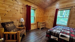 A wooden cabin room with a bed, a chair, a nightstand, and two windows. - Camp Adirondack at Big Moose Lake (Eagle Bay)