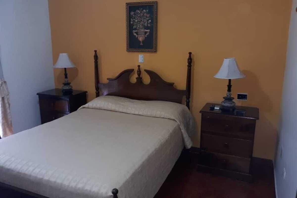 A bedroom with a bed, two bedside tables with lamps, a nightstand, and a wall-mounted picture.
