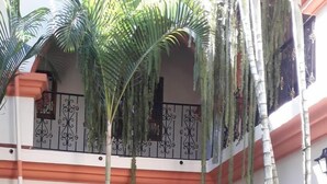 Un patio con un arco central, rodeado de exuberante vegetación tropical y un balcón con barandillas decorativas.