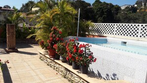 Un área de piscina con suelo de baldosas de piedra, flores rojas en macetas y una valla de celosía blanca.
