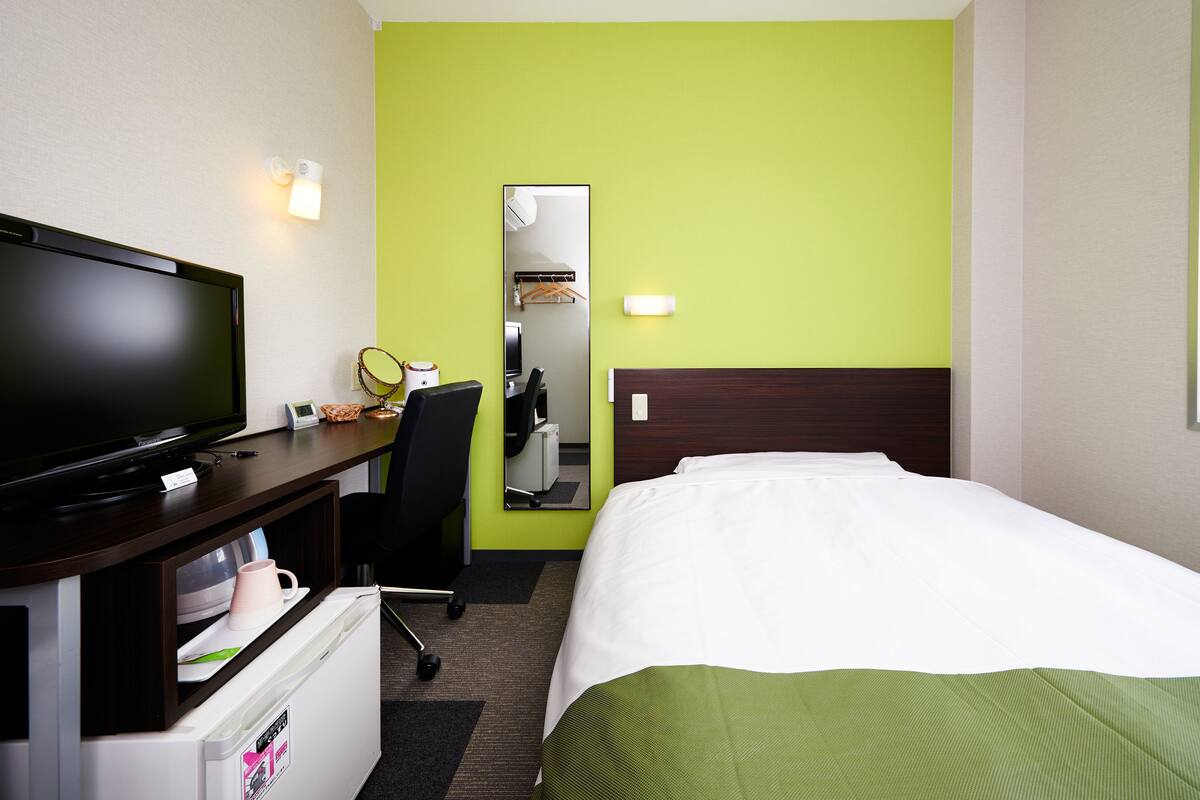 A hotel room with a bed, a desk with a television, a mirror, and a small refrigerator.