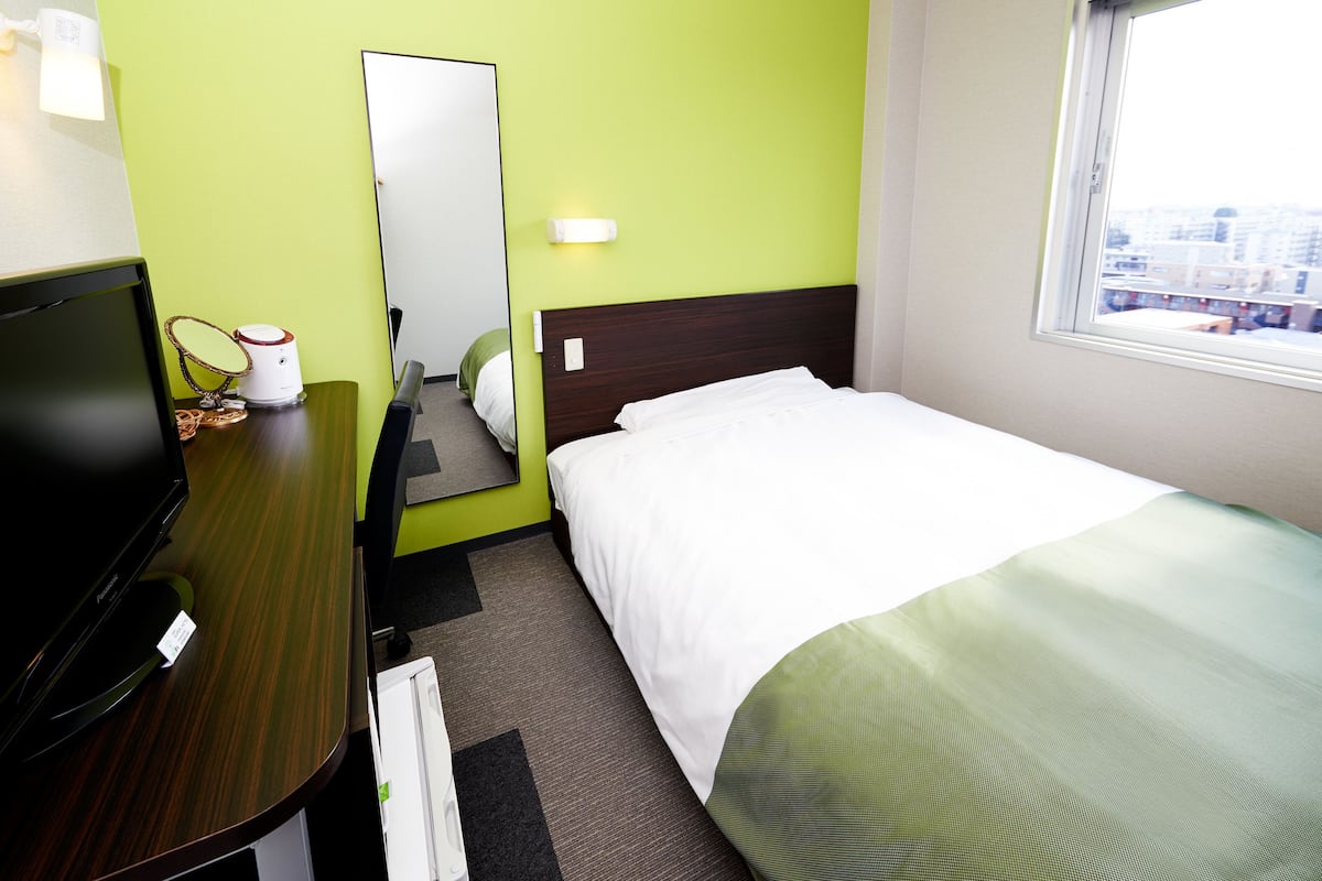 A hotel room with a bed, a desk with a computer, a mirror, and a window with a city view.