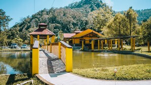 Un puente que lleva a un pabellón amarillo sobre un estanque, rodeado de árboles y colinas.