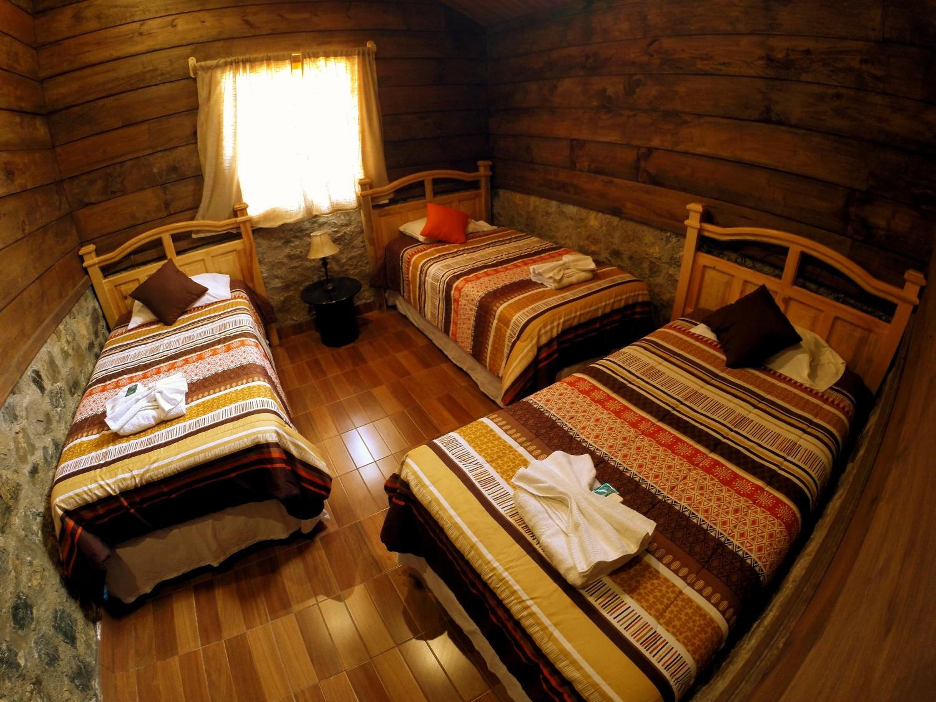 Una habitación de cabaña acogedora con tres camas de madera, cada una con ropa de cama y almohadas estampadas. Una pequeña mesa con una lámpara está entre las camas.