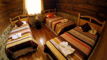 Una habitación de cabaña acogedora con tres camas de madera, cada una con ropa de cama y almohadas estampadas. Una pequeña mesa con una lámpara está entre las camas.