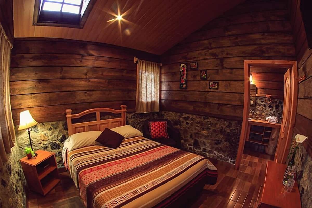 A cozy log cabin bedroom with a bed, a nightstand with a lamp, a stone wall, and a wooden door.
