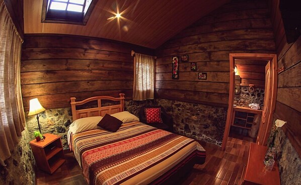 A cozy log cabin bedroom with a bed, a nightstand with a lamp, a stone wall, and a wooden door. - Country Delights Hotel (Purulha)