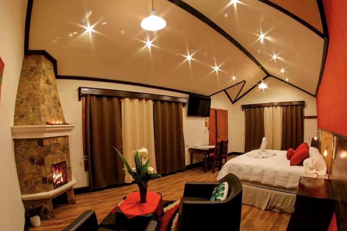 A cozy hotel room with a stone fireplace, a bed with red pillows, and a dining area with a table and chairs.