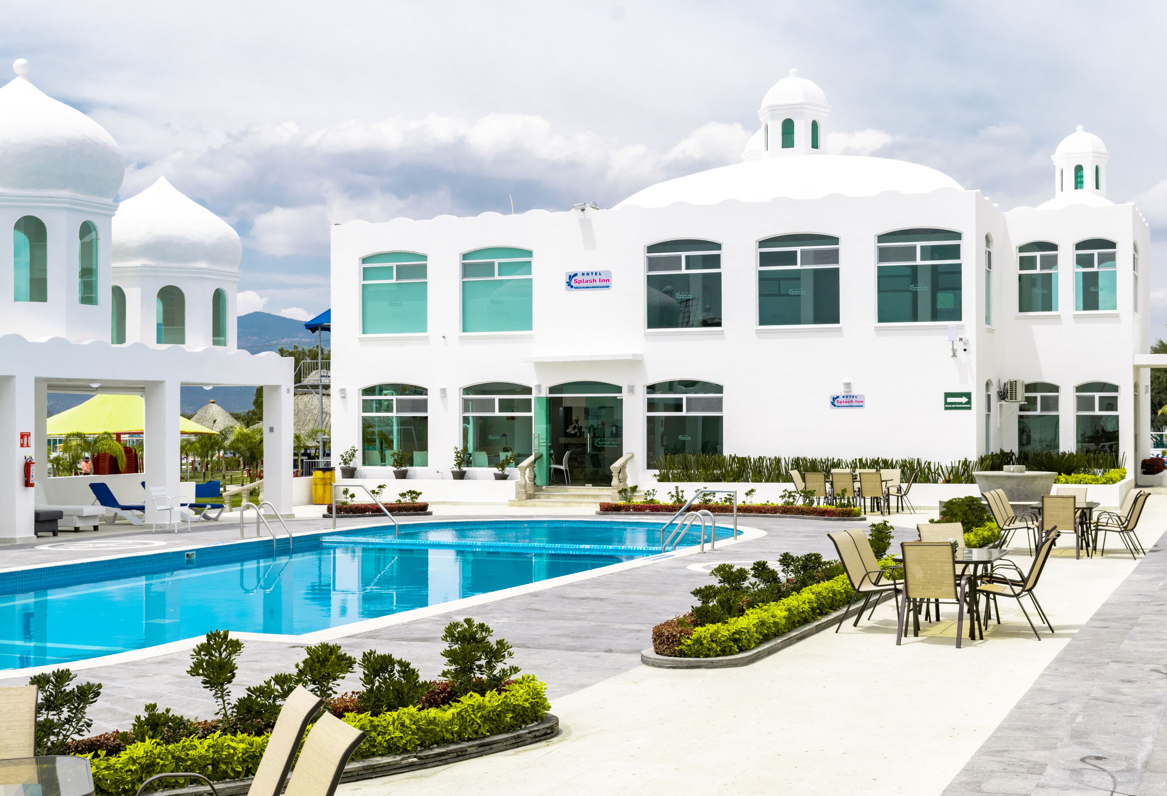 A hotel with a swimming pool, outdoor seating, and a white facade with multiple windows and domed structures.