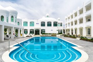 Una piscina con una fuente de agua azul cristalina, rodeada por un edificio blanco con varios balcones y una entrada central.