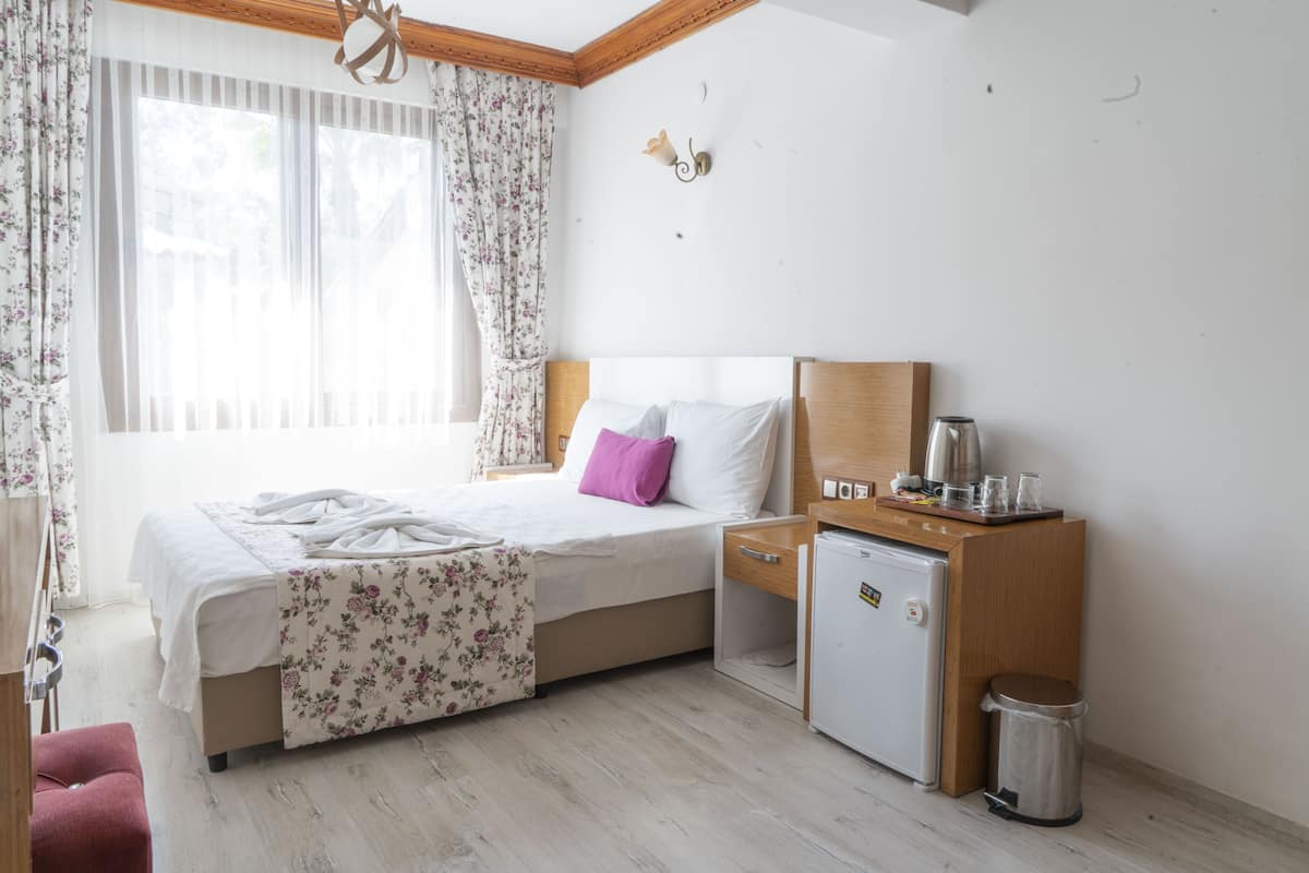 Habitación de hotel con cama, mesita de noche, refrigerador pequeño, cesto de basura y ventana con cortinas.