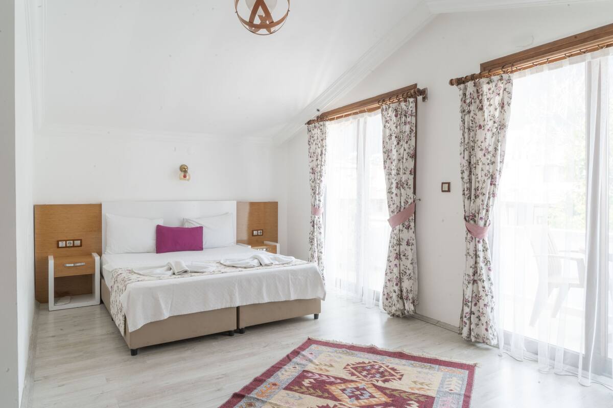 Un dormitorio con cama, mesitas de noche, cabecera de madera, lámpara decorativa en el techo y una ventana con cortinas.