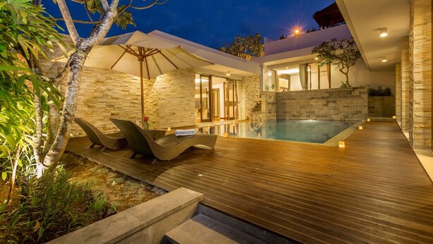 A modern outdoor pool area with stone walls, wooden decking, and lounge chairs. - The Reiko Villa Bali (Nusa Dua)