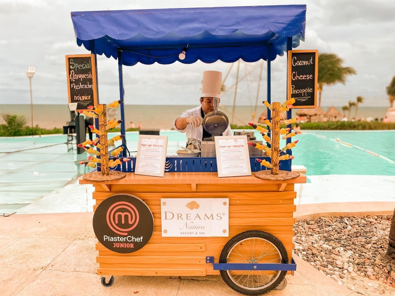 A food cart with a chef, branded with 'PlasterChef Junior' and 'Dreams Natura Resort & Spa' signage, positioned by a poolside.