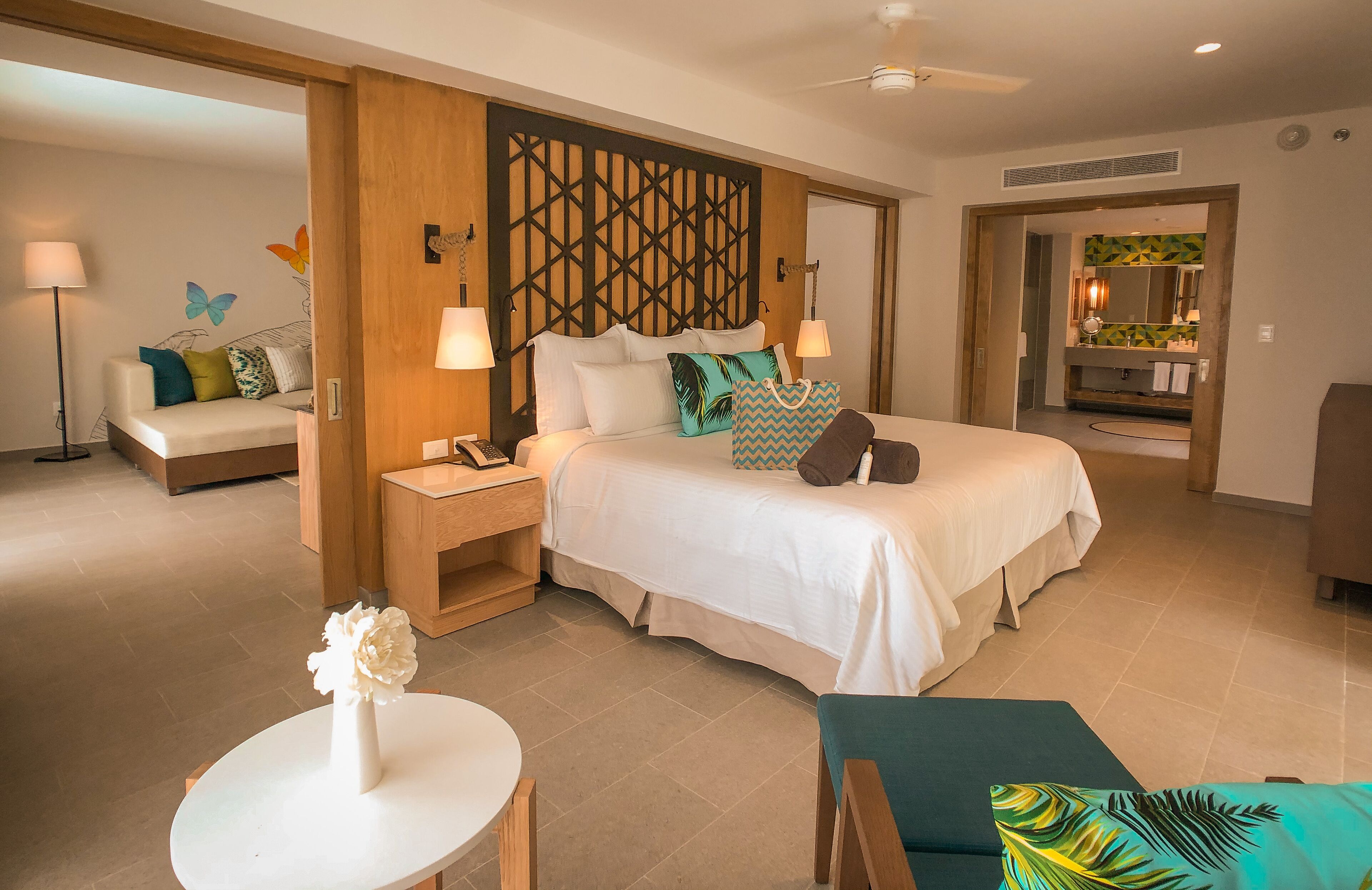Une chambre d’hôtel avec un grand lit, des tables de chevet, un bureau, une chaise, une petite table avec un vase, et une vue sur la salle de bain et la cuisine par une porte ouverte.