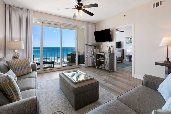 A modern living room with a large window overlooking the ocean, a ceiling fan, a flat-screen TV, and a cozy seating area.