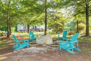 Un espace extérieur aménagé avec quatre fauteuils Adirondack turquoise disposés autour d’un foyer en pierre, le tout entouré d’arbres et avec un bâtiment blanc en arrière-plan.