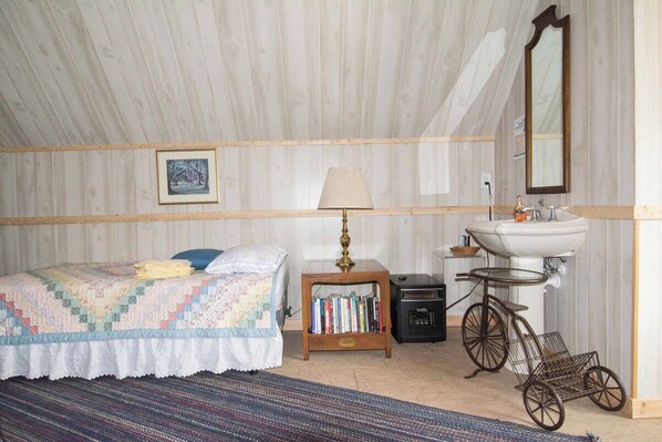 A bedroom with a bed, a bedside lamp, a bookshelf, a vintage bicycle, and a sink.