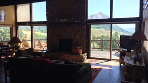 A cozy living room with a stone fireplace, large windows offering mountain views, a comfortable sofa, and a television on a wooden stand. - Rustic mountain home with stunning mountain views from the large deck! (Crested Butte)