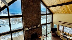 A spacious living room with a stone fireplace, large windows offering a view of a snowy landscape, and a ceiling fan. - Rustic mountain home with stunning mountain views from the large deck! (Crested Butte)