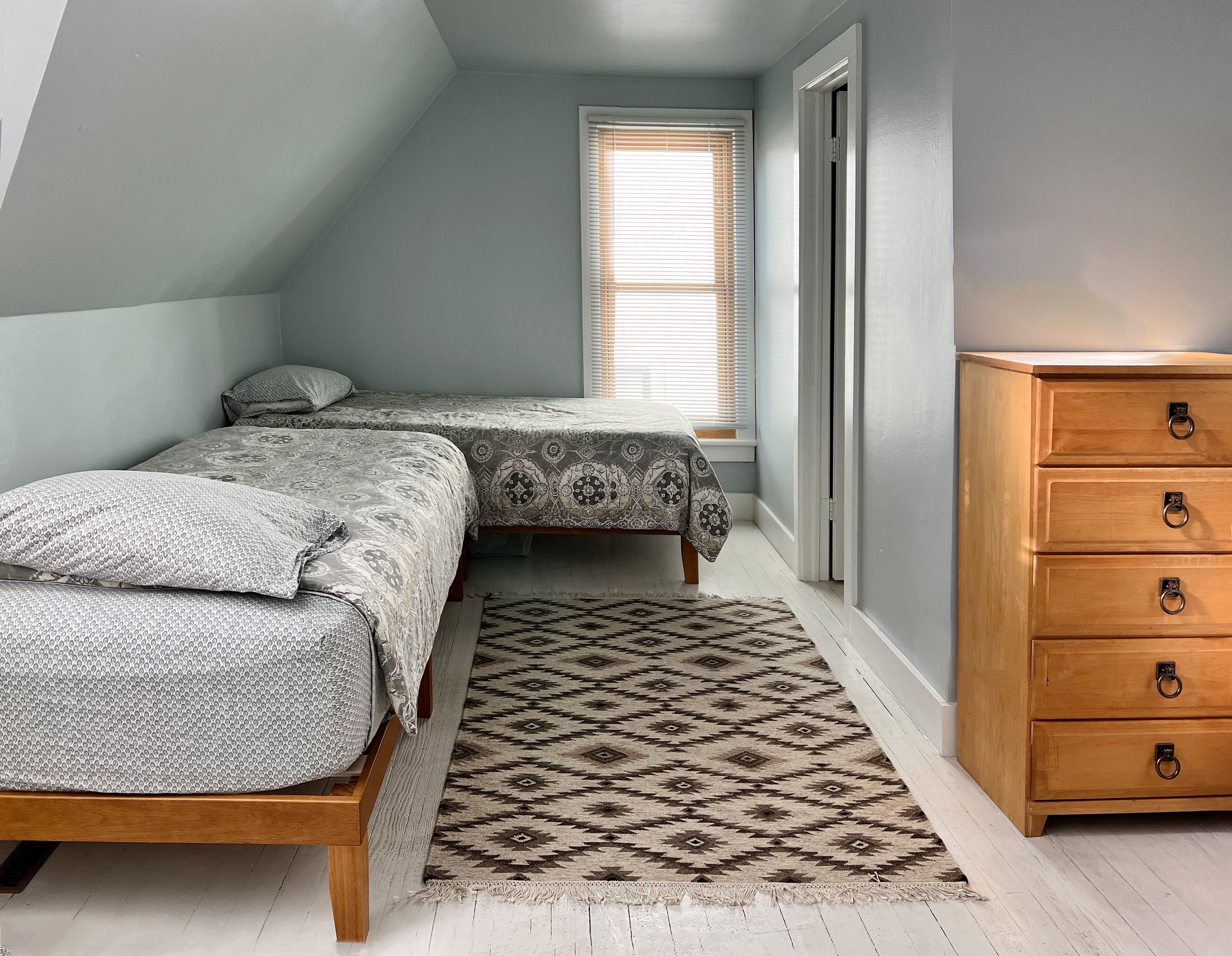 A bedroom with two beds, a wooden dresser, a patterned rug, and a window with blinds.