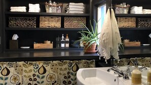 A bathroom with a large white sink, a chandelier, shelves with neatly folded towels, and a patterned curtain.