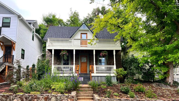 A two-story house with a stone foundation, a front porch with white railings, and a small front yard with steps leading to the entrance.