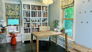 A cozy reading nook with a wooden table, a comfortable chair, a bookshelf filled with books, and a large window with a view of greenery.