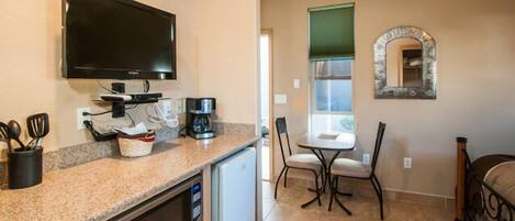 A compact kitchen with a microwave, coffee maker, and a TV mounted above the counter. There is a dining area with a table and chairs, and a small mirror on the wall.