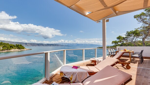 Een zonnig terras buiten met comfortabele zitplaatsen en een tafel, met uitzicht op een heldere blauwe zee en verre bergen.
