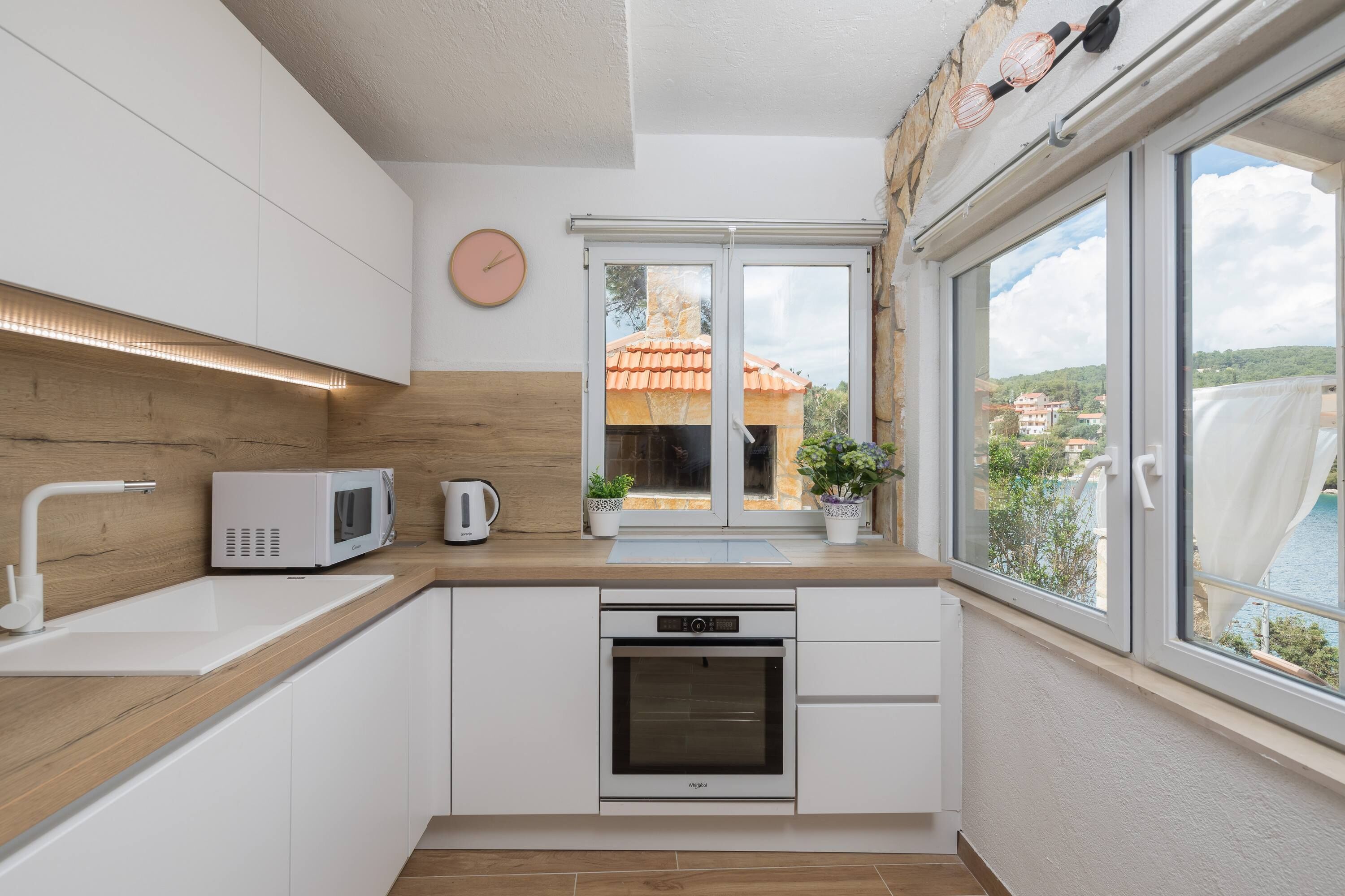 A modern kitchen with white cabinetry, a microwave, a kettle, and an oven. There is a window with a view of a building and a pool.