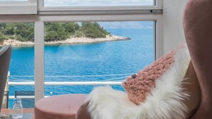 Een kamer met een groot raam met uitzicht op zee en een klein eiland. Er staat een roze stoel met een zacht, wit kussen.