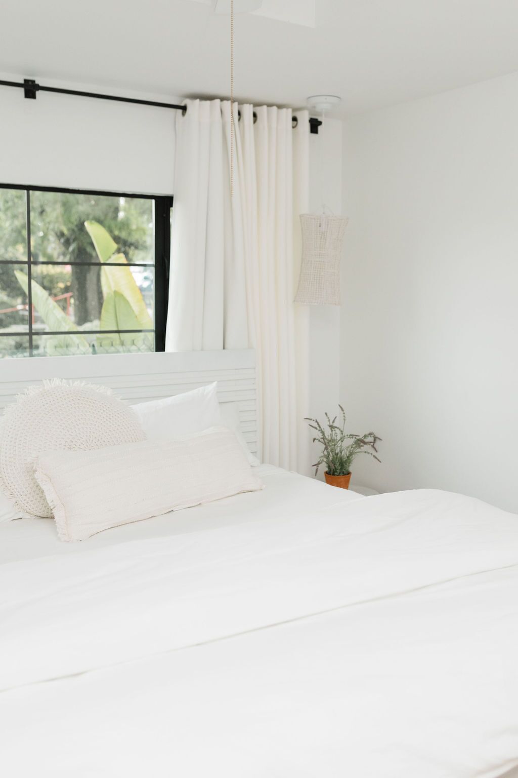 A bedroom with a bed, a window with curtains, a plant, and a wall-mounted light fixture.