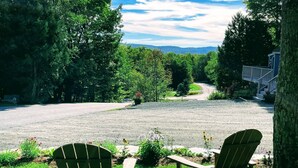 A view of a gravel driveway leading to a house with two Adirondack chairs on a grassy lawn.