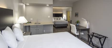 A hotel room with a large bed, a desk with a chair, a small kitchenette, and a separate bedroom with a bed.