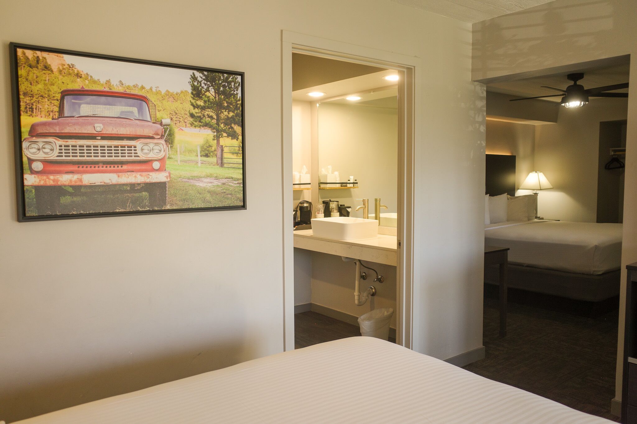 A hotel room with a bed, a bathroom with a sink and mirror, and a painting of a vintage car on the wall.