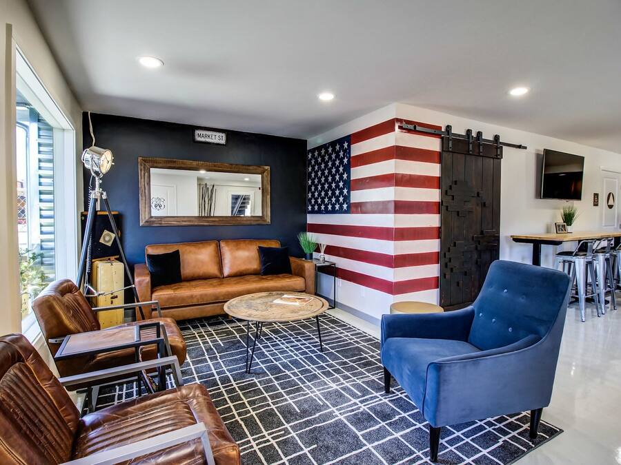 Een moderne woonkamer met een leren bank, een blauwe fauteuil en een salontafel. Een muurschildering van de Amerikaanse vlag siert de wand, en een schuifdeur leidt naar de keuken.