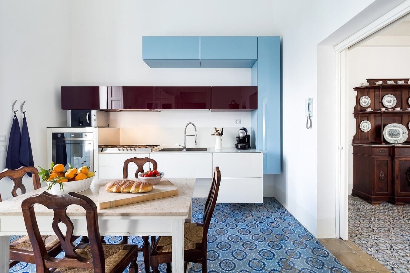A compact kitchen with white cabinets, a blue upper section, a dining area with a marble table, and patterned floor tiles.
