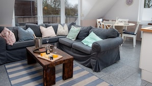 Ein modernes Wohnzimmer mit einer grauen Eckcouch, einem Holztisch und einem Streifenmuster-Teppich. Es gibt einen Essbereich mit einem weißen Tisch und Stühlen, sowie eine Küchenzeile mit einem weißen Schrank und einer Uhr an der Wand.